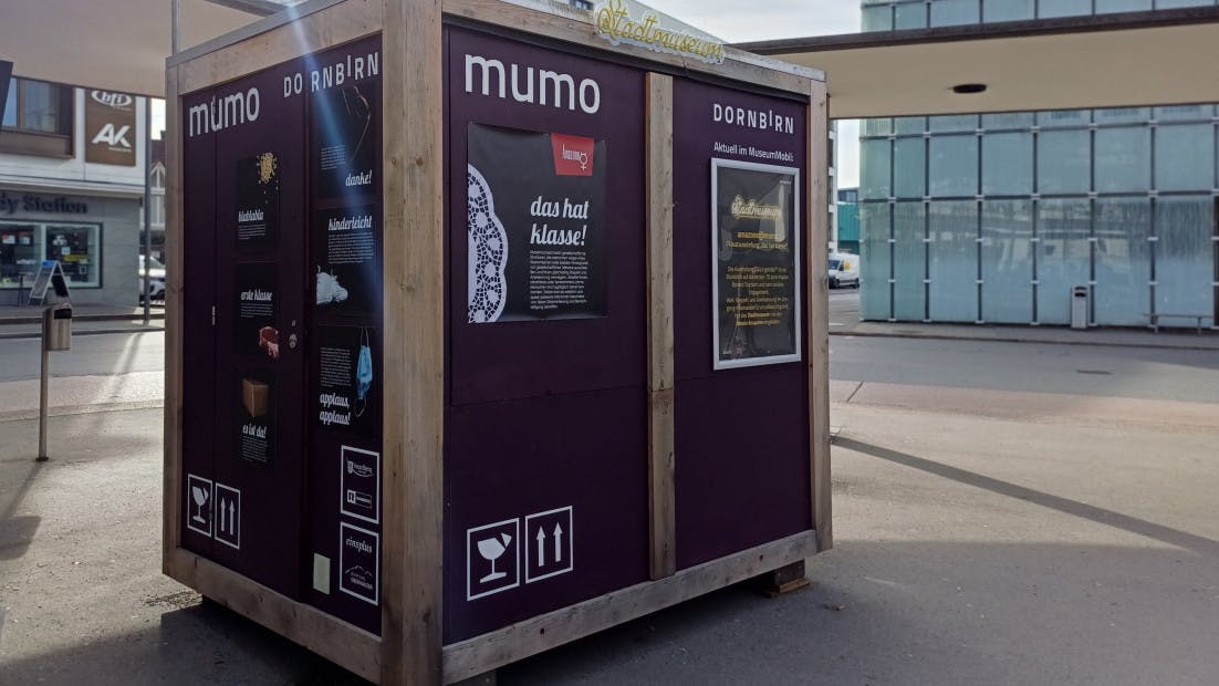 Holzkubus mit der Beschriftung mumo ist mit Plakaten versehen und steht auf einem öffentlichen Platz.